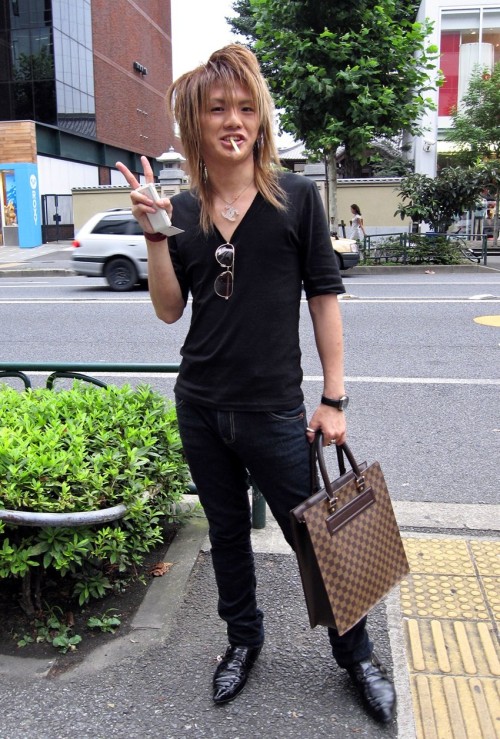 A fashionable Japanese guy posing on the street in Harajuku while smoking. He&#8217;s wearing a black v-neck shirt, black denim jeans, pointy black leather shoes, and a Chanel necklace. He also has a checkered brown leather handbag, sunglasses hanging from his shirt, several rings, and a white mobile phone in his hands.