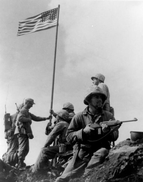 A US Marine keeps watch with his M1A1 carbine during the first flag raising on Iwo Jima, 1945